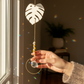 Hand holding a monstera leaf shaped suncatcher near a window.