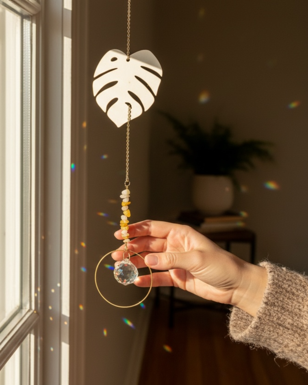 Hand holding a monstera leaf shaped suncatcher near a window.